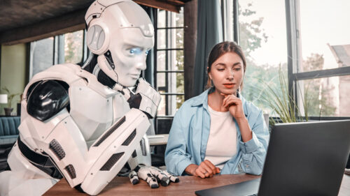 Ein Roboter und eine Frau sitzen gemeinsam vor einem Laptop an einem Tisch.