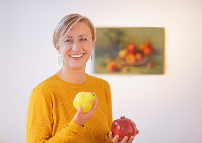 Blonde Frau mit gelbem Pullover hält zwei Früchte in ihren Händen und lächelt in die Kamera. Im Hintergrund ist verschwommen ein Gemälde mit Obstkorb zu erkennen.