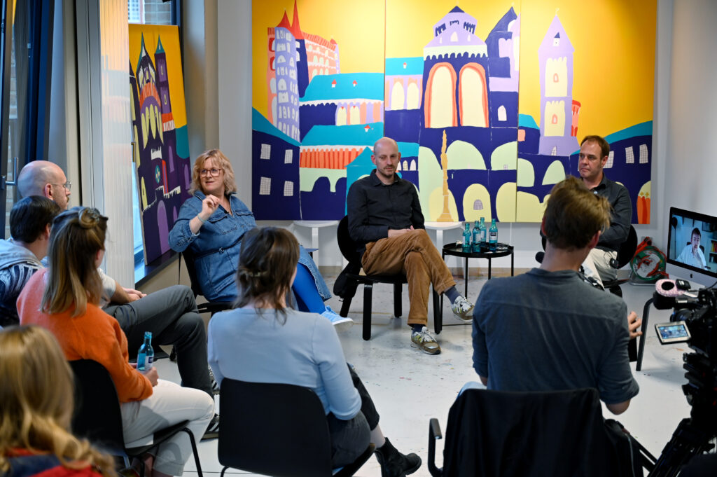 Gruppendiskussion in einem modernen Raum. Die Teilnehmenden sitzen in einem Halbkreis auf Stühlen. Im Hintergrund sind bunte Wandgemälde und Kunstwerke zu sehen. Kameras und Beleuchtungsausrüstung deuten auf eine Filmproduktion hin. Der Raum hat große Fenster mit Blick auf ein Stadtgebiet. Die Atmosphäre wirkt entspannt und konzentriert, mit verschiedenen Personen unterschiedlichen Alters, die aufmerksam zuzuhören oder zu diskutieren.