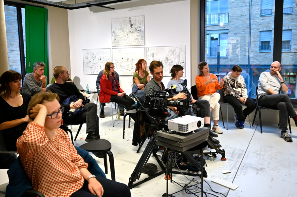 Gruppendiskussion in einem modernen Raum. Die Teilnehmenden sitzen in einem Halbkreis auf Stühlen. Im Hintergrund sind bunte Wandgemälde und Kunstwerke zu sehen. Kameras und Beleuchtungsausrüstung deuten auf eine Filmproduktion hin. Der Raum hat große Fenster mit Blick auf ein Stadtgebiet. Die Atmosphäre wirkt entspannt und konzentriert, mit verschiedenen Personen unterschiedlichen Alters, die aufmerksam zuzuhören oder zu diskutieren.